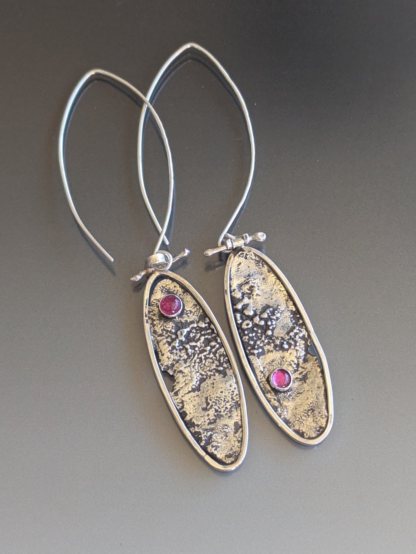 Silver oval earrings with textured design and ruby gemstones on a gray background
