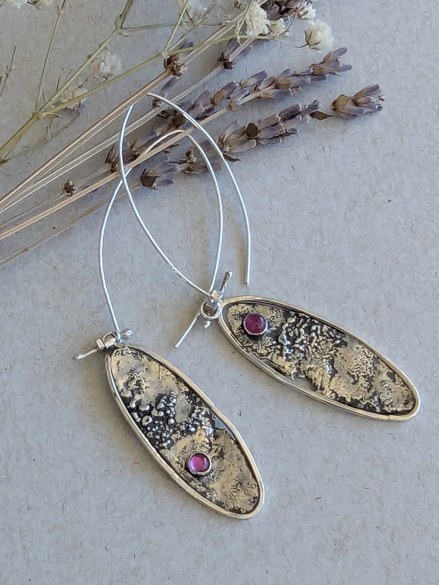 Pair of oval silver earrings with textured accents and ruby gemstones on a light background.