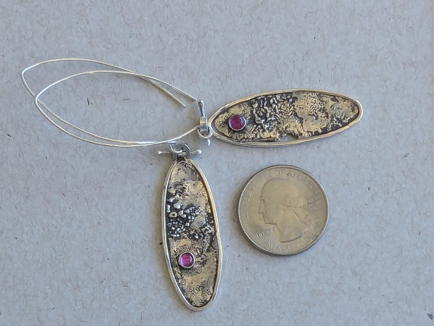 Silver oval earrings with textured design and ruby stones, next to a quarter for scale on a gray background