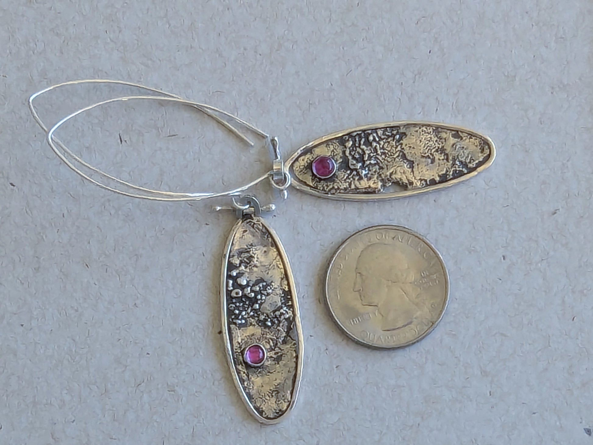 Silver oval earrings with textured design and ruby stones, next to a quarter for scale on a gray background