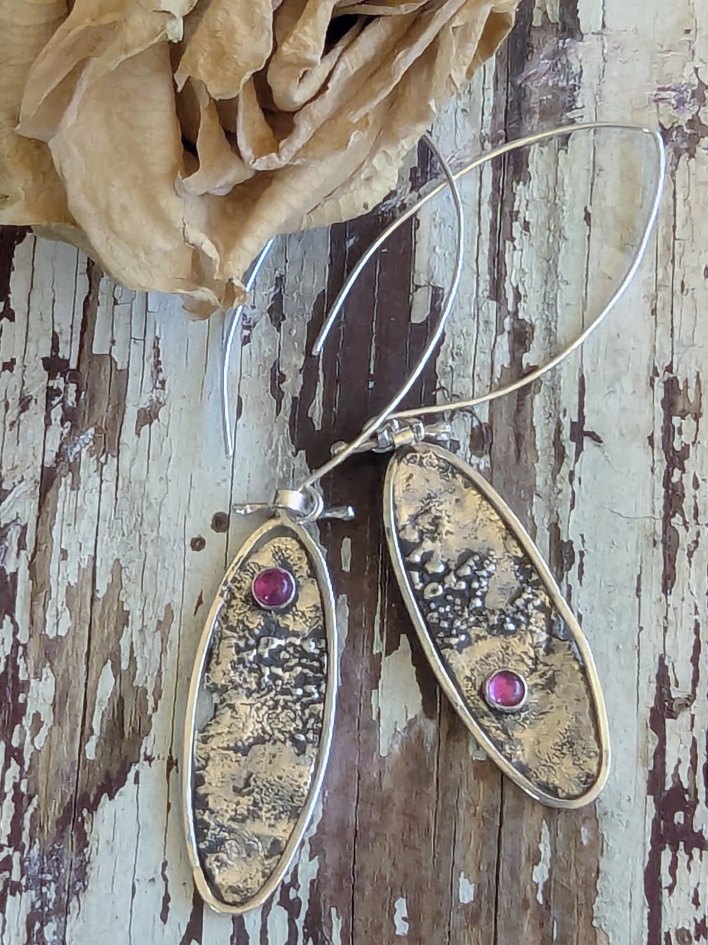 Pair of silver oval earrings with textured design and ruby gemstones on a wooden surface