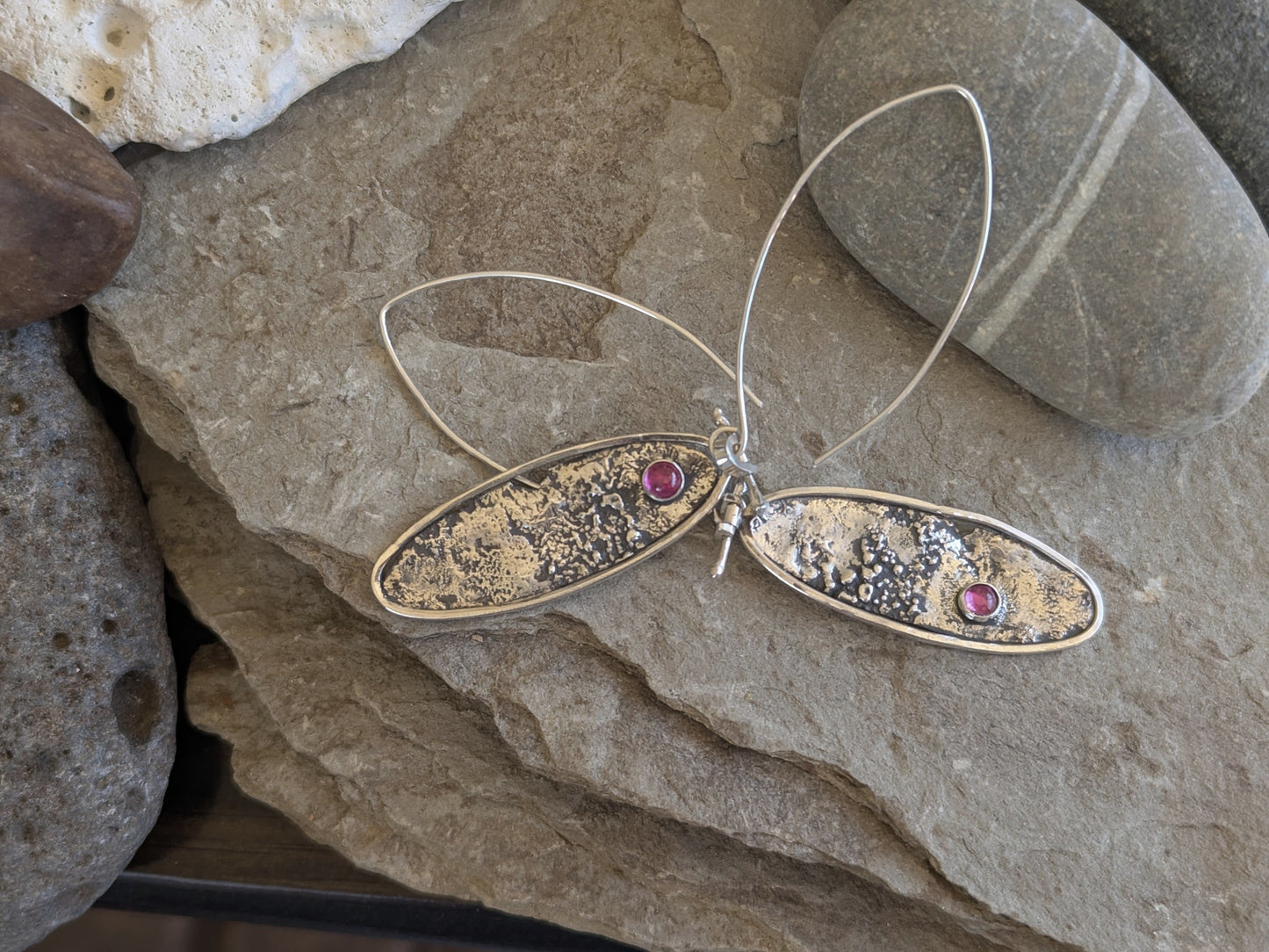 Silver oval earrings with textured design and ruby gemstones on a stone surface
