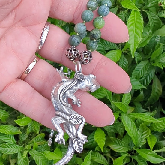 video of hand holding silver lizard pendant on green bead and silver necklace in front of leafy background
