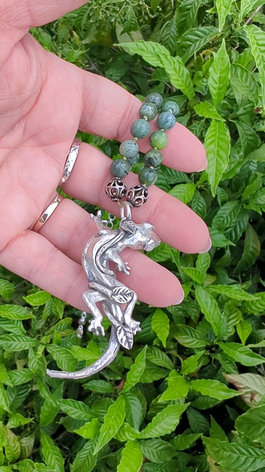 video of hand holding silver lizard pendant on green bead and silver necklace in front of leafy background